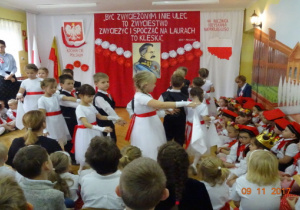 Na tle biało czerwonej dekoracji tańczą w parach odświętnie ubrane dzieci.